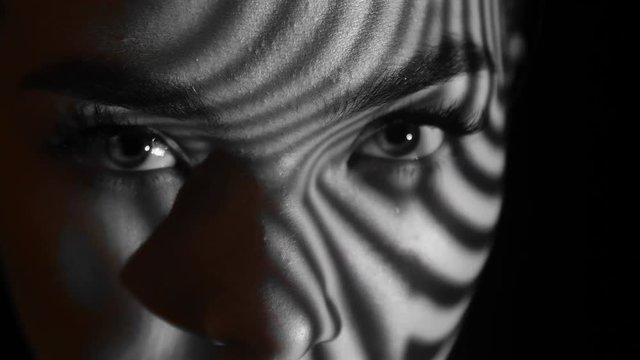 Projection of black and white lines on girl’s face. Close up eyes of girl with an abstract pattern of lines and red lips on her face. Illumination from the blinds in the room. Concept. Sharp look.
