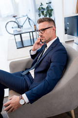 businessman in glasses and suit sitting in armchair and talking on smartphone