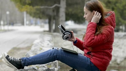 Close-up, the teenager takes off his headphones and looks at the digital tablet screen, sitting on the rocks in the city spring Park in a red jacket and with red hair on his head. - Powered by Adobe