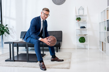 cheerful businessman in suit holding basketball in hands while sitting on coffee table - Powered by Adobe