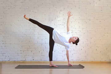 The girl is a professional instructor of hatha yoga practicing asanas in the room against the background of a white brick wall. Ardha Chandrasana (Half Moon Pose).