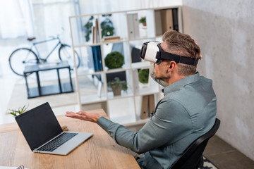 businessman wearing virtual reality headset while sitting near laptop with blank screen