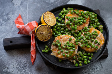 Cast-iron pan with roasted hake medallions, green peas and lemon, horizontal shot on a grey concrete background
