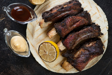Barbecued beef ribs with dipping sauces served on tortillas, view from above, dark brown stone background