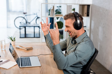 happy businessman listening music in headphones and looking at laptop while showing rock sign in office