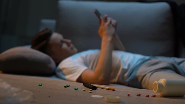 Addicted teenager scrolling phone, medication capsules and cigarette on table