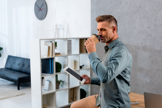 handsome businessman drinking coffee drom disposable cup and holding digital tablet in office