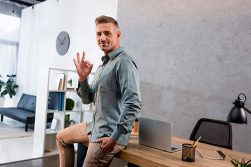 happy businessman showing ok sign while sitting with hand in pocket in office