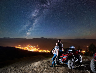 Active man and woman trevelers with atv quad motorbike on the top of mountain, romantic couple enjoying beautiful view of night sky full of stars, Milky way, luminous town on background © anatoliy_gleb