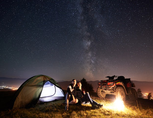 Young couple man and woman trevelers sitting near glowing tourist tent, burning campfire, atv quad motorbike on the top of mountain, enjoying beautiful view of night sky full of stars and Milky way © anatoliy_gleb