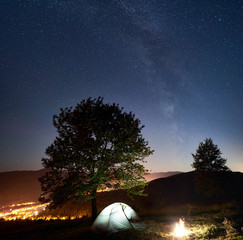 Tourist camping near big tree at night. Glowing tent and bonfire under magical night sky full of stars and Milky way. On background incredibly beautiful starry sky, mountains and luminous town