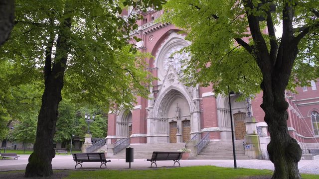 Static shot of the main entrance, to St Johns Church, filmed under green leaves and branches, waving in the wind, on a cloudy, summer day, in Ullanlinna, Helsinki, Finland