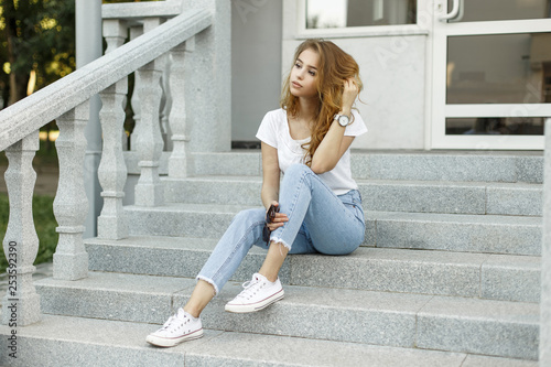 Modern Attractive Stylish Young Woman In A Fashionable T Shirt In Blue Jeans In White Sneakers Resting Sitting On The Steps Of A Vintage Building Cute European Girl Enjoying Summer Day Outdoors Modern Attractive Stylish Young Woman In A Fashionable T Shirt In Blue Jeans In White Sneakers Resting Sitting On The Steps Of A Vintage Building Cute European Girl Enjoying Summer Day Outdoors