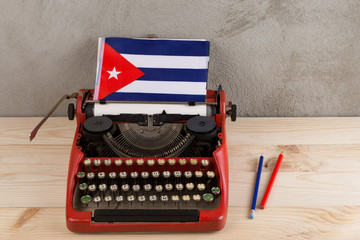 Political, news and education concept - red typewriter, flag of the Cuba, pencils on gray cement background