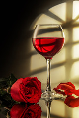 a glass of red wine and a red rose on the table with reflection, light from the window