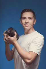 Smiling Man with camera isolated on blue background. Young man holding digital camera and making photo looking on you. Lifestyle, travel and technology concept. Happy boy in white t-shirt with dslr