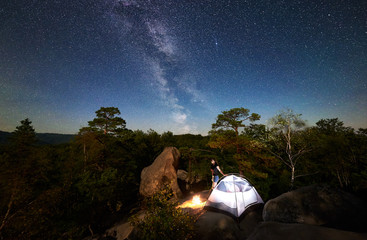 Woman traveller relaxing on rocky mountain top beside camp, bonfire and tourist tent at summer night, enjoying view of sky full of stars and Milky way. On background starry sky, big boulders and trees