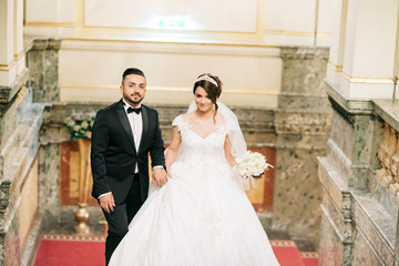 A beautiful young Turkish man wearing a black suit and a beard standing in the home along with his beautiful bride in a white dress and a bouquet of flowers