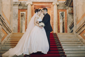 Obraz premium A beautiful young Turkish man wearing a black suit and a beard standing in the home along with his beautiful bride in a white dress and a bouquet of flowers