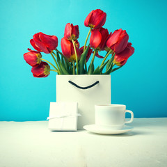 Bouquet of brightly red tulips in a white gift bag, white cup with coffee, white gift box on a blue background. Concept congratulations, surprises and gifts