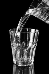 Water pouring into the glass from measuring cup on black reflective background.