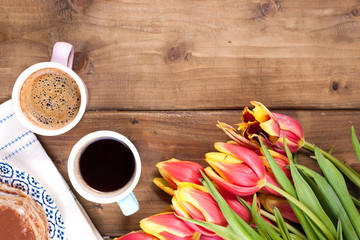 Two cups of aromatic coffee and pancakes with cream. Breakfast for two on a wooden background and a bouquet of red spring tulips. Free space for text.