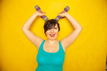 chubby joyful emotional girl with short hair in a blue t-shirt doing sports with dumbbells to be healthy and lose weight on yellow solid studio background