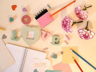Decorative composition for congratulations on Mother's Day from fresh pink carnations, the volumetric letters, hearts, empty notebooks, crayons, paper, pencils on a light background, top view