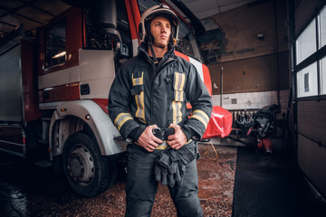 Portrait of a brave young fireman wearing protective uniform standing next to a fire engine in a garage of a fire department