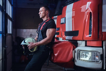 Fototapeta na wymiar Muscular fireman holding a protective helmet in a garage of a fire department, leaning on a fire engine and looking outside