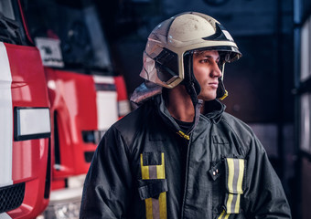 Fototapeta premium Brave young fireman wearing protective uniform standing next to a fire engine in a garage of a fire department and looking outside