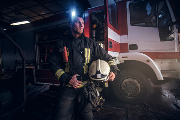 Fototapeta na wymiar Fireman wearing uniform holding a helmet and looking at a camera while standing near a fire truck in a garage of a fire department at night.
