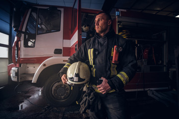 Obraz na płótnie Canvas Handsome fireman wearing uniform holding a helmet and looking sideways while standing near a fire truck in a garage of a fire department
