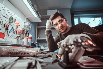 Stylish electronic technician in goggles, leaning on a desk in a repair shop, looking at a camera...