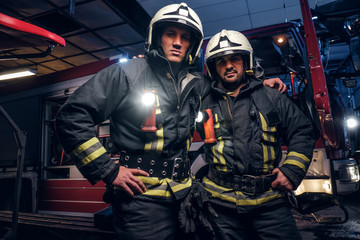 Two firemen hug and looking at the camera standing near the fire truck at night