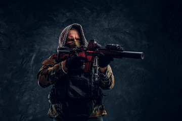 Special forces soldier in military uniform wearing mask and hood holding an assault rifle with a laser sight and aims at the target. Studio photo against a dark textured wall