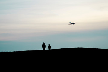 Fototapeta na wymiar The silhouettes of two people stand on a high hill and watch an airplane flying high in the sky