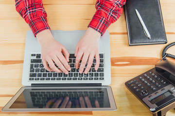 Top view of a stylish wooden office desk with a keyboard, a gray laptop and notebook and pen, working mans hands.