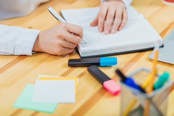 Office table with accessories: a marker of blue and pink colors, a pen, a pen, a pencil, a notebook. During this hand, the young woman begins to write a pen in a notebook.