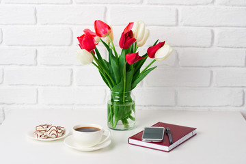 tulip flowers are in a vase on the table, cup of coffee, diary and smartphone, white brick wall as background