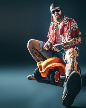 Cooler, tätowierter Mann mit Bart auf Spielzeugauto / Kurze Hose, Hemd und Sonnebrille