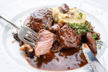 Traditionell geschmorte Schweinebäckchen in brauner Rotwein Sauce mit Pilzen und Kartoffel Püree als closeup auf einem klassischen weißen Design Teller