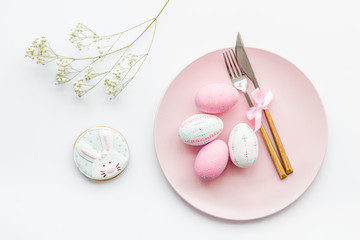 Easter dining table in pastel colors. Plate, cutlery, painted eggs, gingerbread and dry flower branch on white background top view