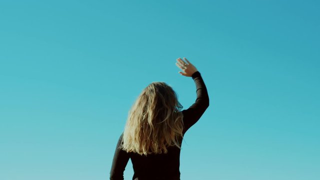 Simple clean shot with copy space of young woman with blonde hair wave goodbye or hello to imaginary plane person in distance on beautiful blue or turquoise sky. Concept greeting and holiday mode