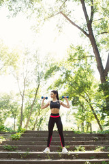 Young woman exercising with dumbbells at the park. Female fitness model working out outdoor. Concept of healthy lifestyle.	