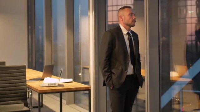 Front view of successful businessman in suit standing in office with hands in pockets