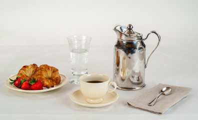 Cream Colored Plate and Coffee Cup with Two Croissants and Three Fresh Strawberries on a White Linen Tablecloth and a Silver Hotel Room Service Carafe with a Napkin and Spoon.