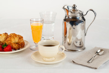 Cream Colored Plate and Coffee Cup with Two Croissants and Three Fresh Strawberries and Orange Juice on a White Linen Tablecloth and a Silver Hotel Room Service Carafe with a Napkin and Spoon.