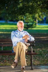 Fototapeta premium Senior man relaxing in park on a sunny day seated on a wooden bench and waiting for someone