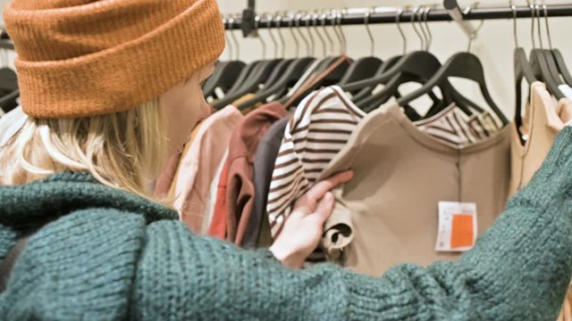 A girl in a green sweater and a yellow hat walks through a store of things and chooses what to buy. Touches things on hangers and looks at price tags